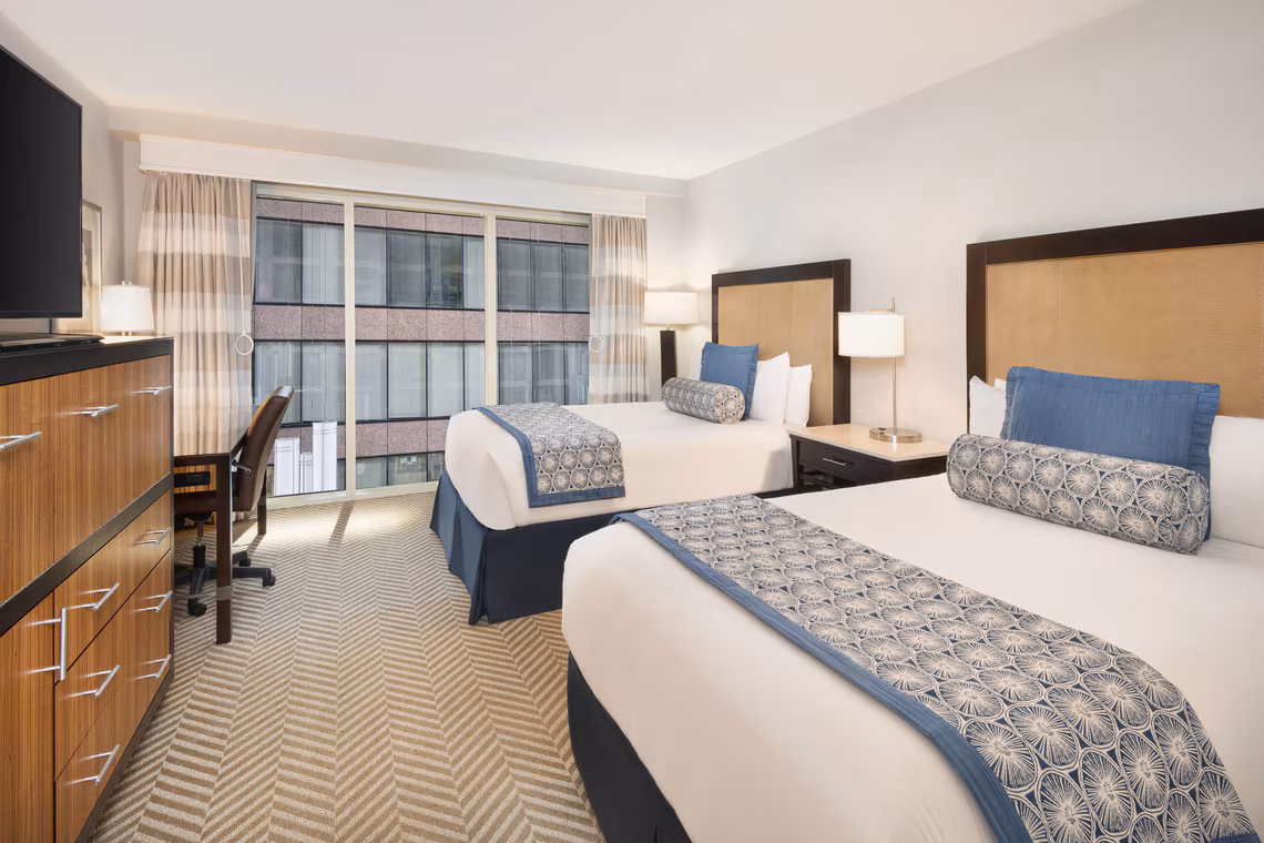 Hotel room with two double beds, blue and white bedding, desk, TV, lamps, dresser, and large windows with city view.
