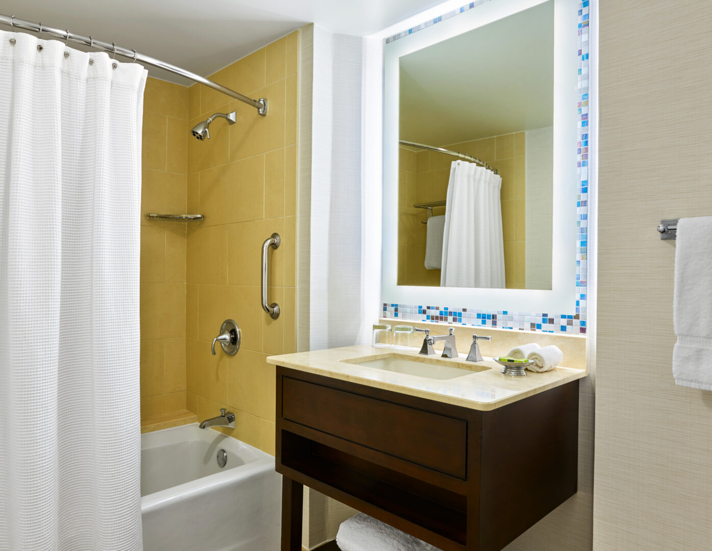 Bathroom with yellow tiled bathtub, white shower curtain, grab bar, dark wood sink cabinet, large lit mirror, and towel hanging on wall.