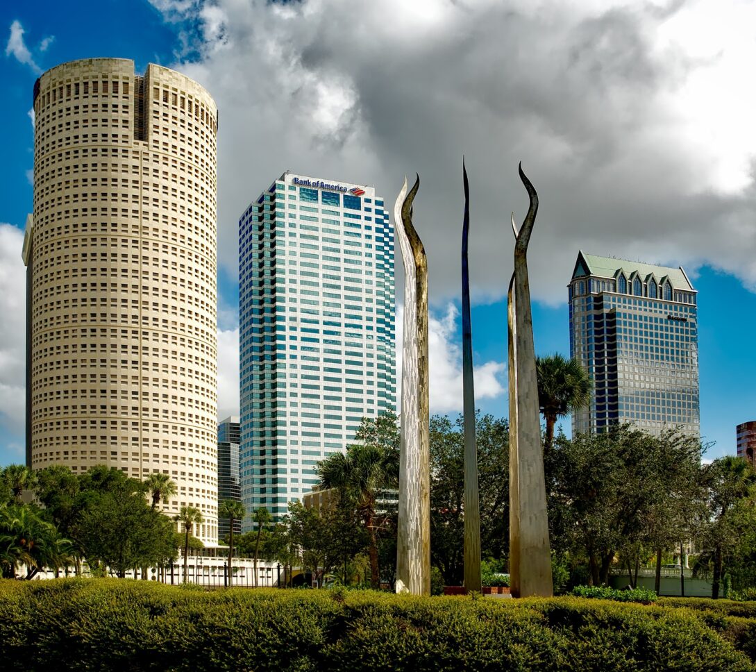 Three tall metal sculptures stand before modern skyscrapers and palm trees under a partly cloudy blue sky.