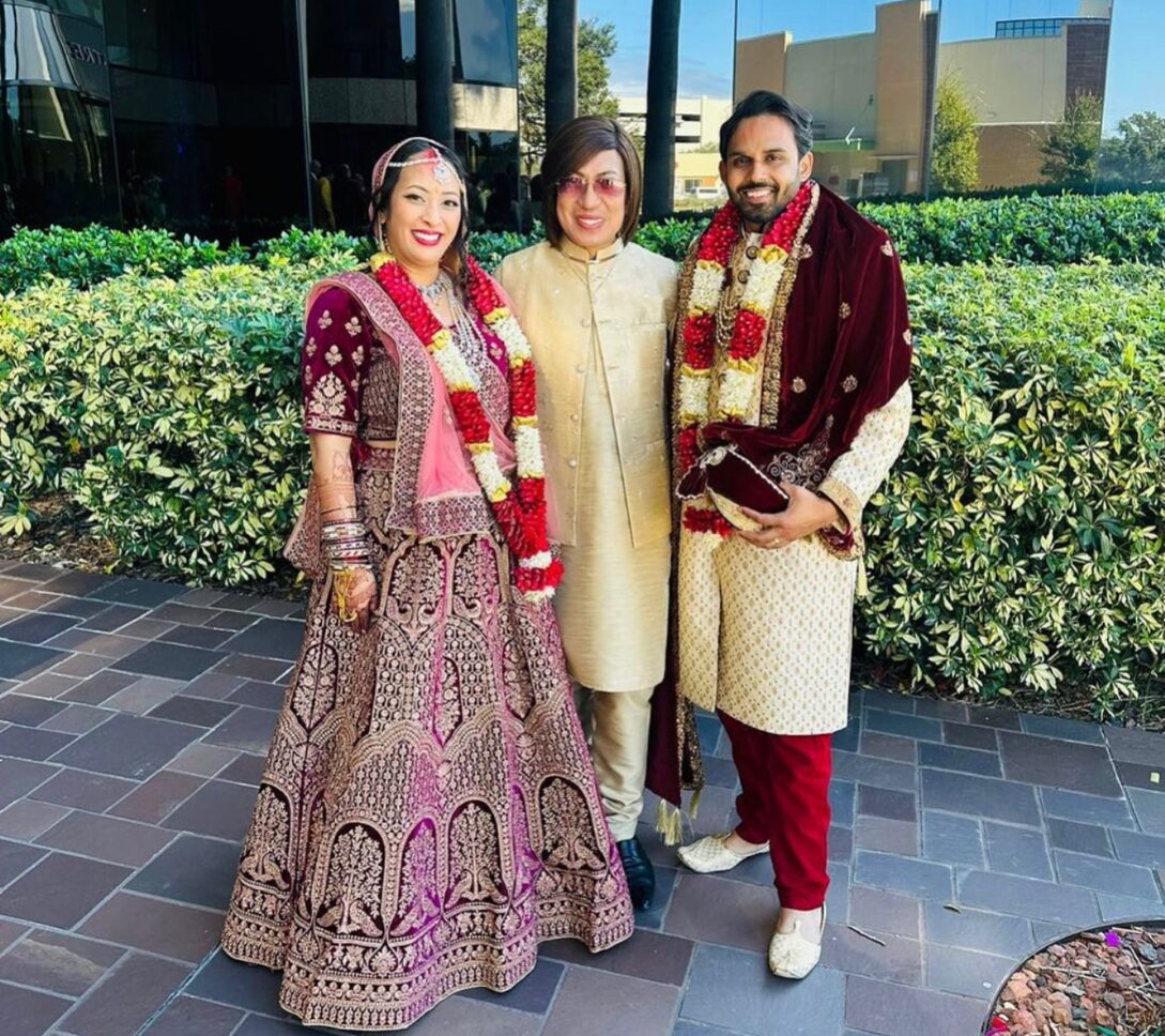 Three people in traditional Indian attire pose and smile outdoors in front of a glass building and greenery; two appear to be a bride and groom.