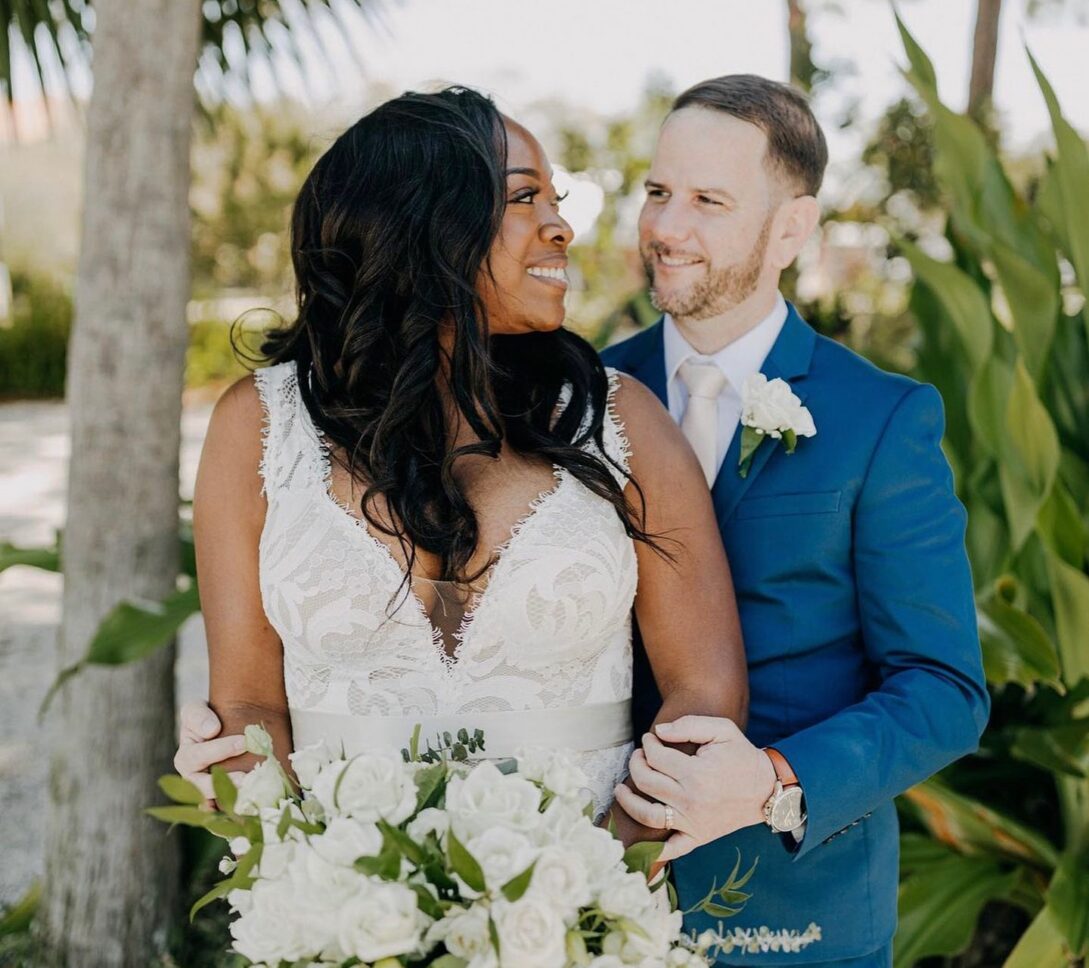 Bride in a white dress holding white roses, groom in blue suit standing behind her, both smiling outdoors among green foliage.