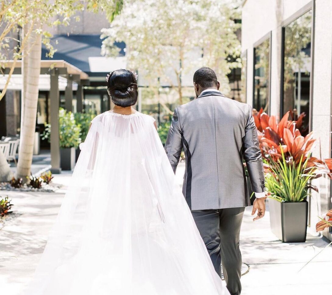 A bride in a flowing white gown and a groom in a gray suit walk hand in hand outdoors, surrounded by plants and sunlight.