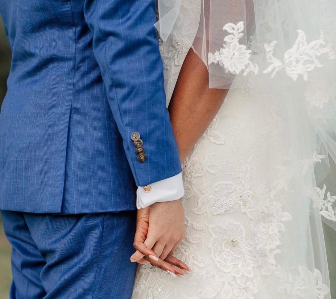 A bride in a lace wedding dress and a groom in a blue suit hold hands closely, with part of her veil and dress details visible.