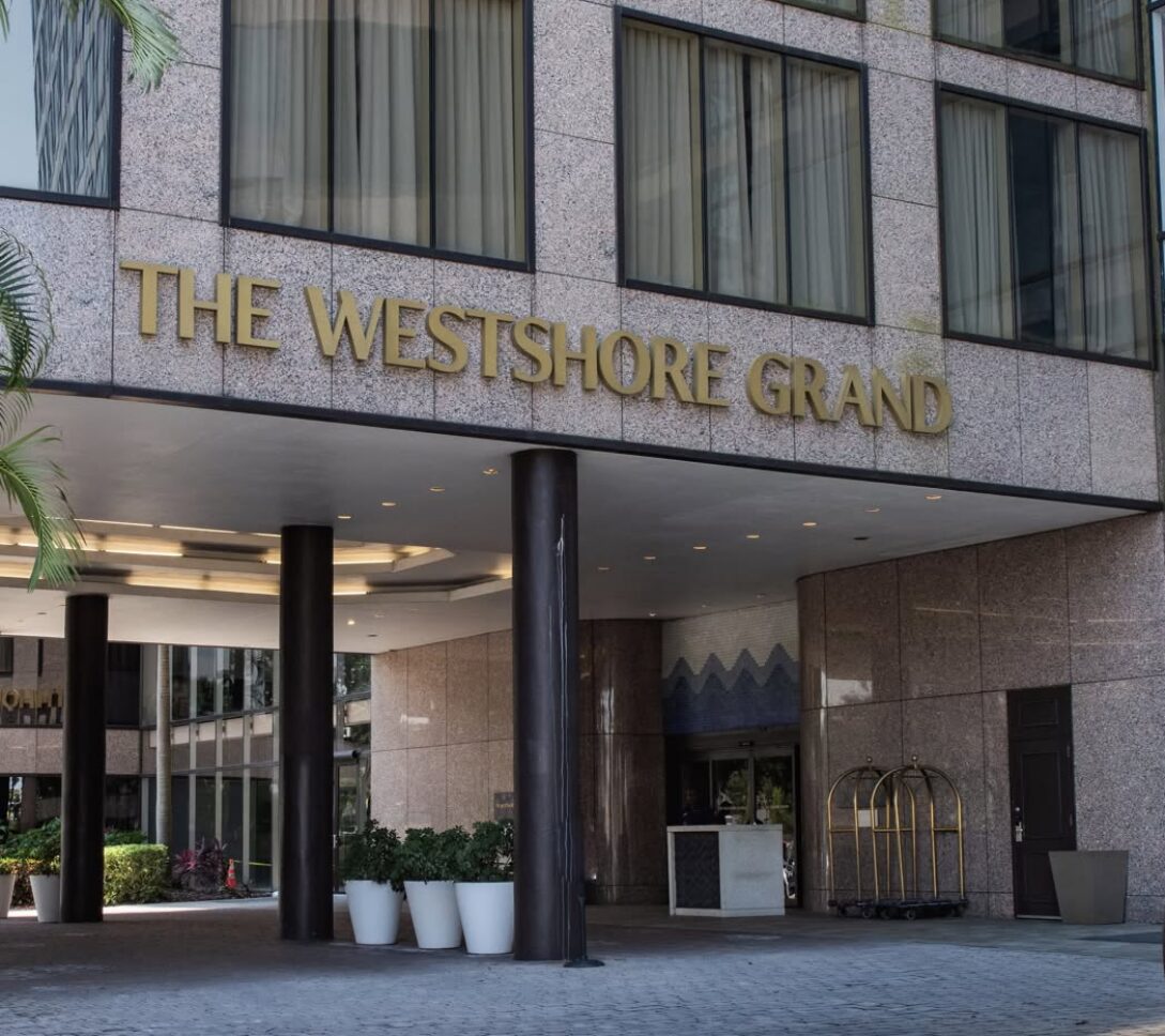 Entrance of The Westshore Grand hotel, featuring large gold signage, glass windows, potted plants, and a luggage cart near the doorway.