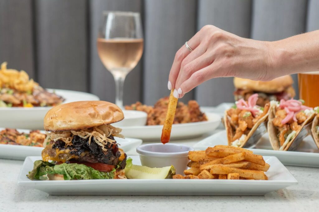 A hand dips a seasoned French fry into sauce, next to a burger with lettuce and tomato, more fries, and tacos in the background.