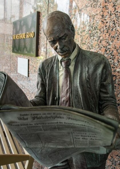 A realistic bronze statue of a man in a suit reading a newspaper stands by benches outside the Westshore Grand Hotel.