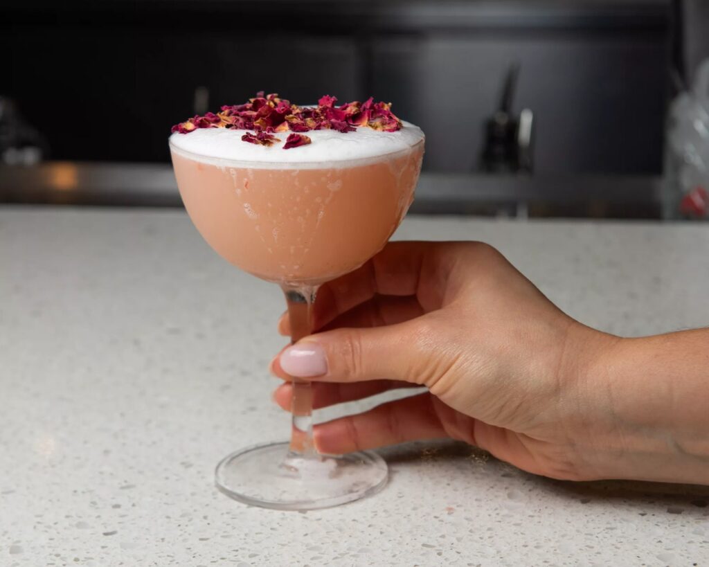 A hand holding a pink cocktail in a coupe glass, topped with foam and dried rose petals, on a speckled countertop.