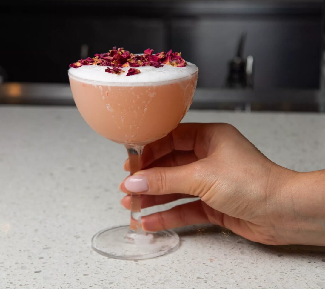 A hand holding a pink cocktail in a coupe glass, topped with foam and dried rose petals, on a speckled countertop.
