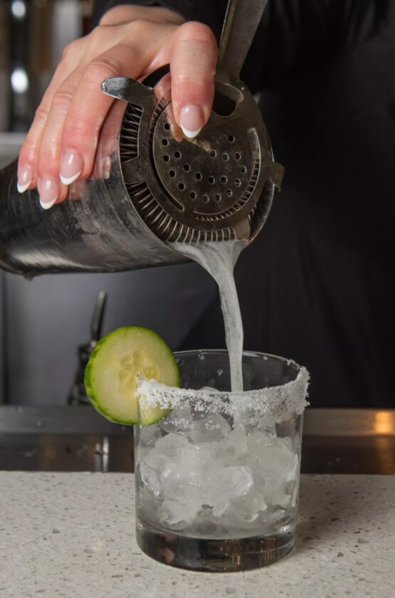 A hand pours a cocktail from a shaker into a salt-rimmed glass with ice and a cucumber slice on the rim, set on a bar counter.