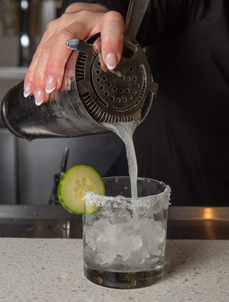 A hand pours a cocktail from a shaker into a salt-rimmed glass with ice and a cucumber slice on the rim, set on a bar counter.