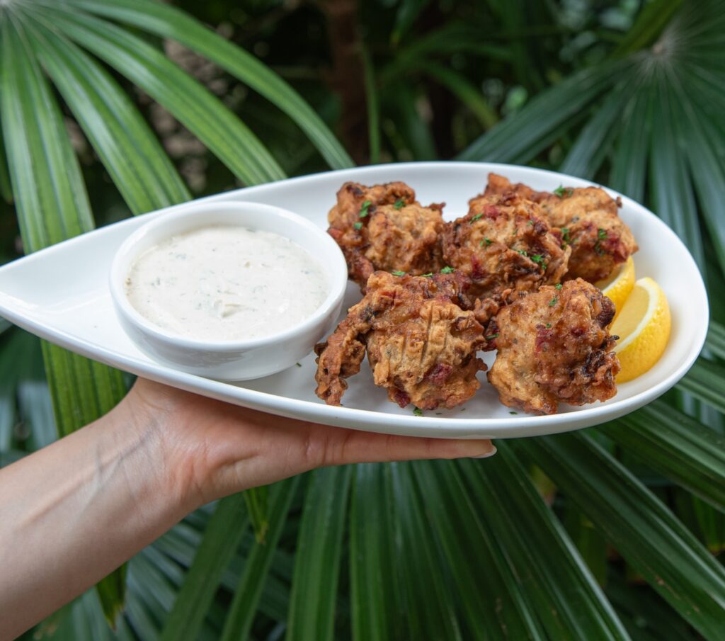 A hand holds a plate with four fritters, lemon wedges, and a bowl of dipping sauce, in front of green palm leaves.