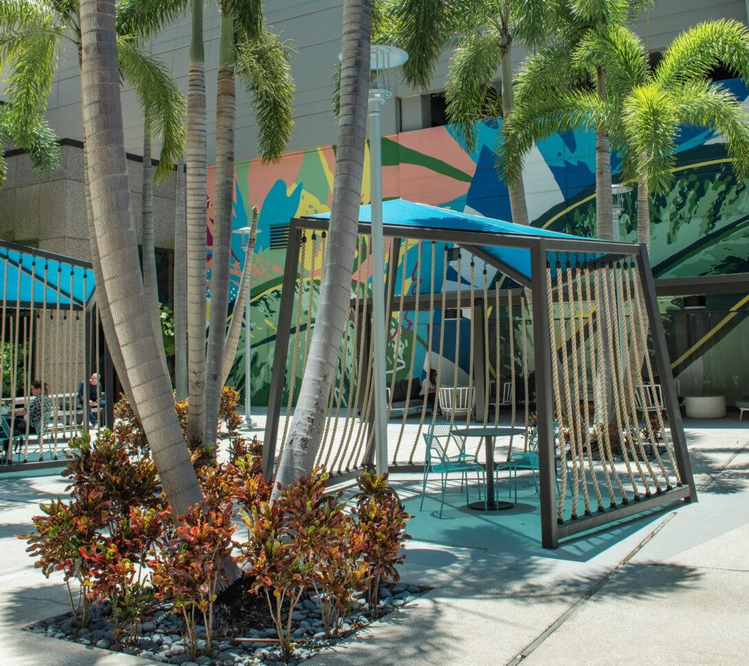 Tall palm trees and colorful plants surround modern swings and chairs in an outdoor plaza, with a vibrant mural and parking garage in the background.