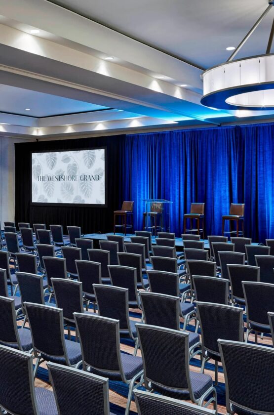 Rows of empty chairs face a stage with three chairs and two screens at the Westshore Grand Hotel’s modern conference room with blue lighting.
