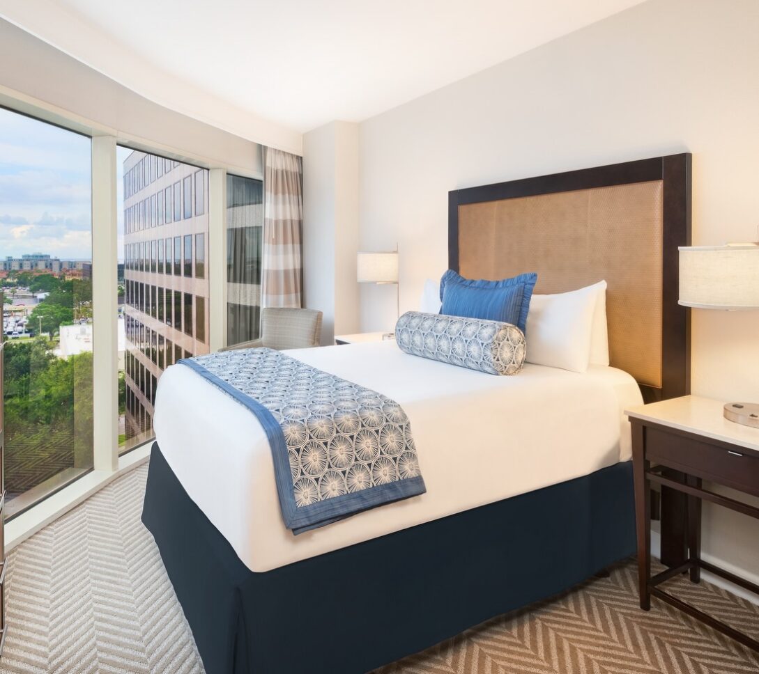 A hotel room with a neatly made bed, a large window with a city view, modern furniture, and neutral decor with blue accents.
