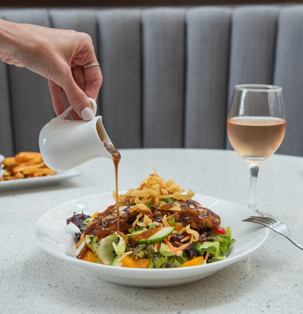 A hand pours dressing over a salad with fried onions. A glass of rosé wine and fries are on the table.