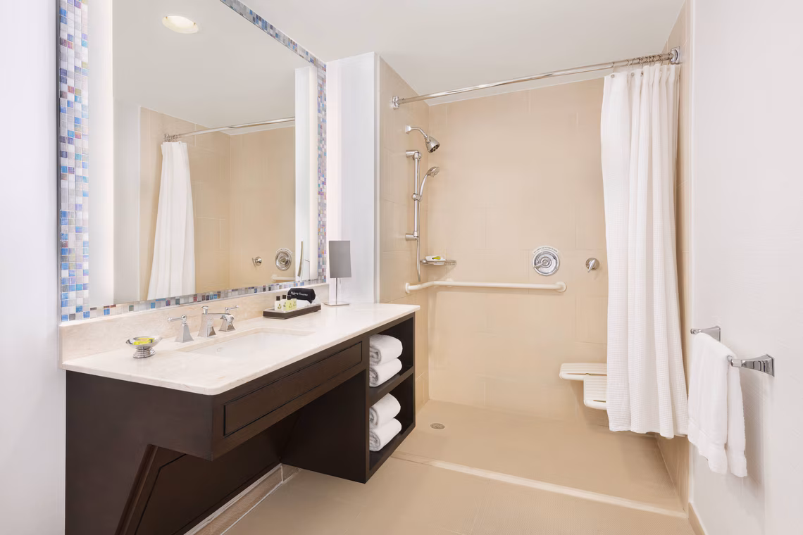 Accessible bathroom with roll-in shower, grab bars, handheld showerhead, fold-down seat, large vanity, mirror, towels, and white curtains.