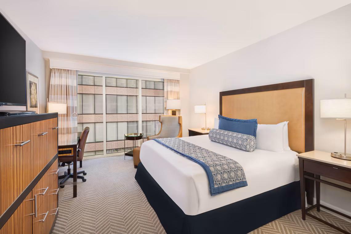Hotel room with a king bed, blue accents, work desk, armchair, lamps, TV, and large window with city view.