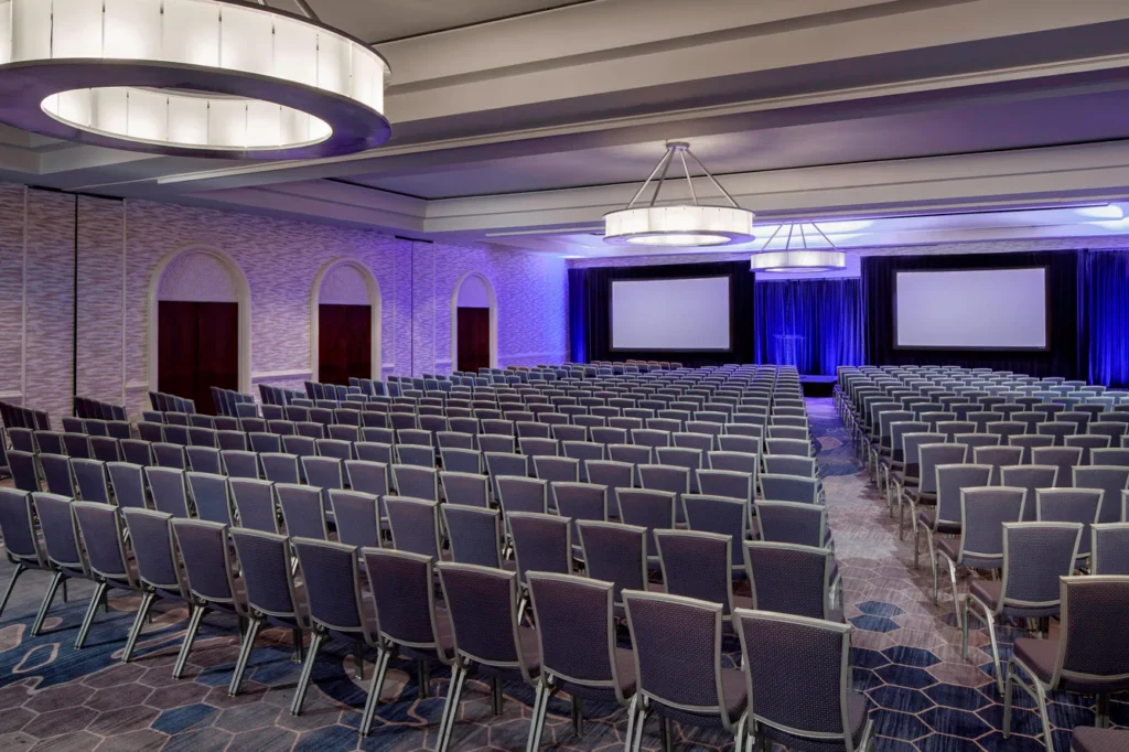 Rows of gray chairs face two large projection screens in a modern conference room with purple lighting and geometric carpet patterns.