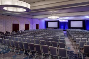 Rows of gray chairs face two large projection screens in a modern conference room with purple lighting and geometric carpet patterns.