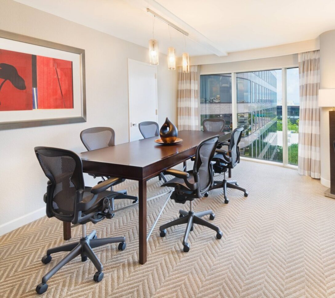 Modern meeting room with six chairs around a table, large windows with city views, a desk, artwork on the wall, and neutral décor.