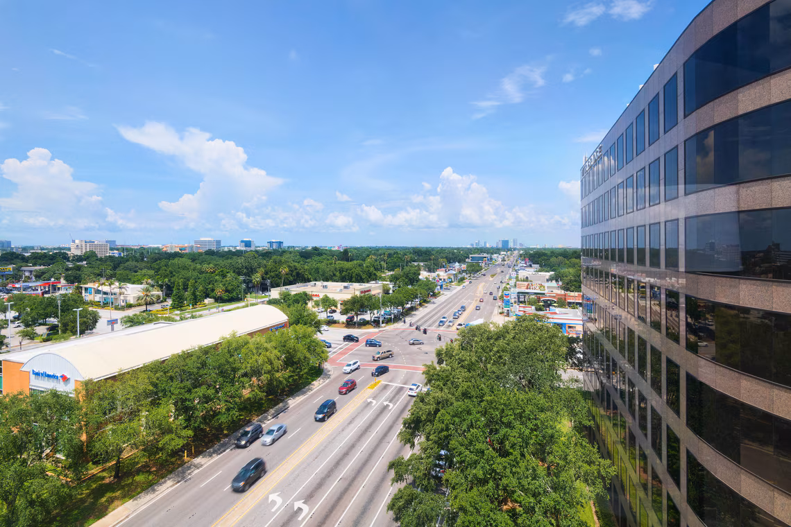A busy city street with cars, surrounded by trees and buildings, under a blue sky with scattered clouds.