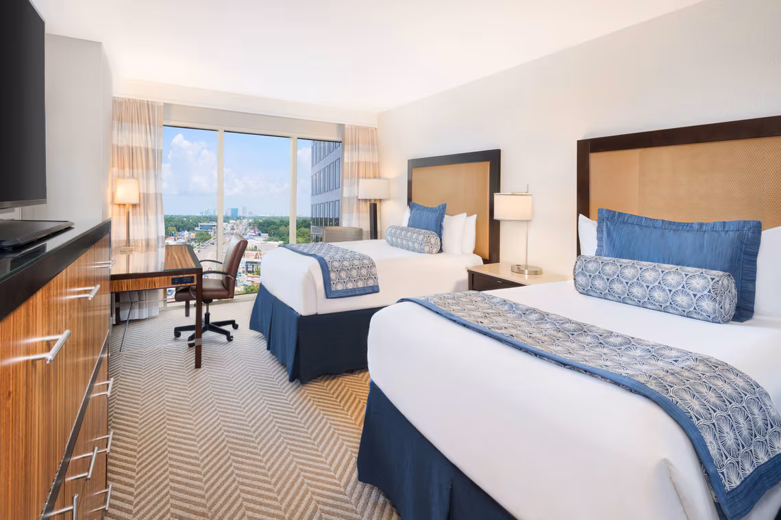Hotel room with two double beds, blue and white bedding, desk with chair by a large window, and TV on a wooden dresser. City view outside.