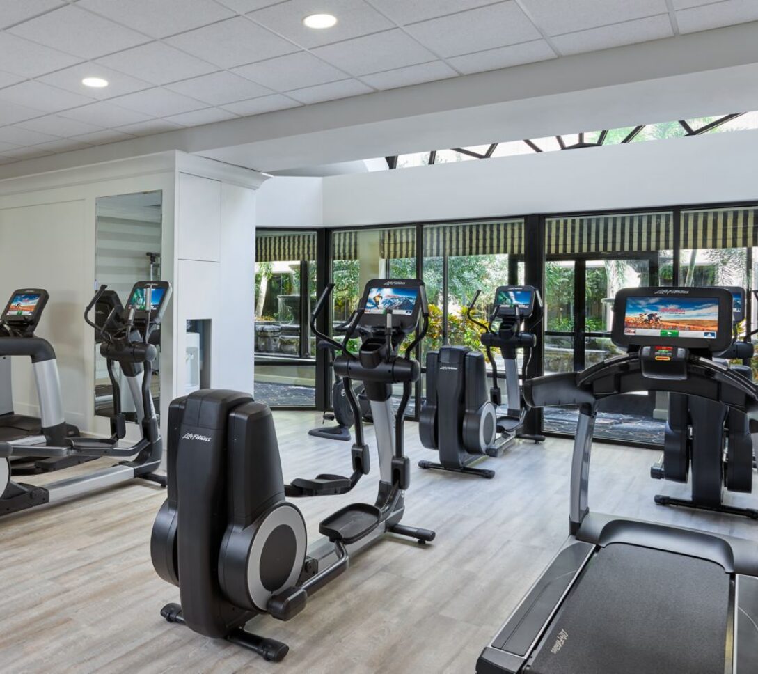 Modern gym with treadmills, ellipticals, and stationary bikes. Large windows let in natural light and offer a view of greenery outside.