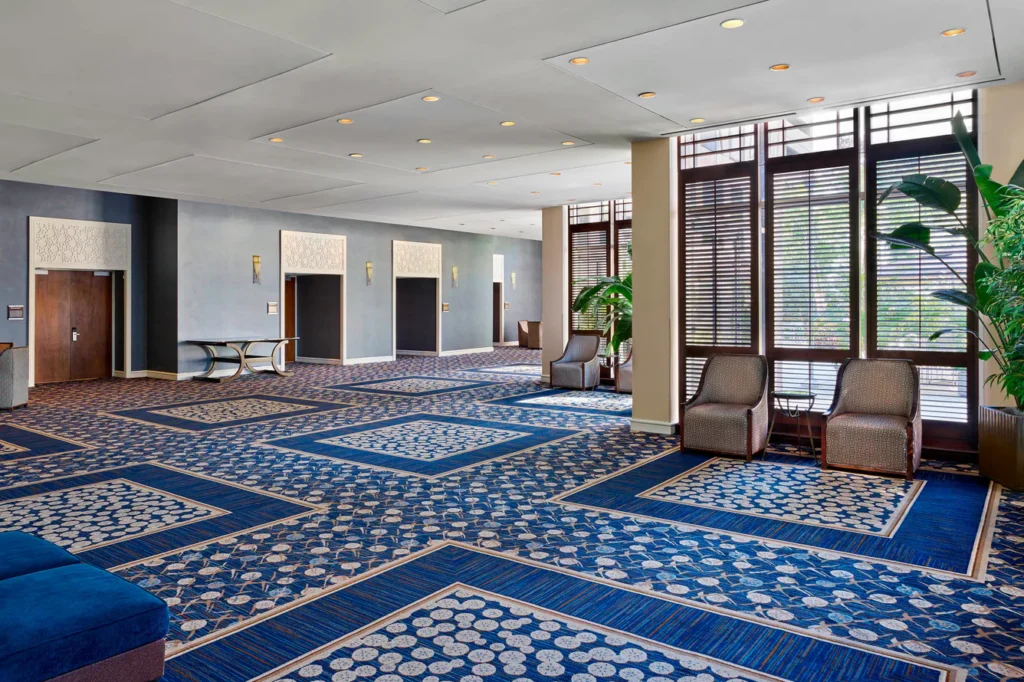 Spacious hotel lobby with blue patterned carpet, large windows with blinds, armchairs, plants, and elevators along the back wall.
