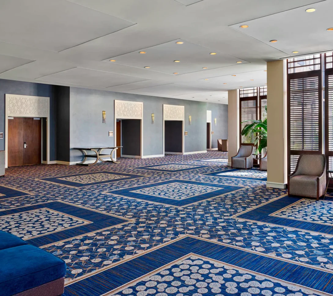 Spacious hotel lobby with blue patterned carpet, large windows with blinds, armchairs, plants, and elevators along the back wall.
