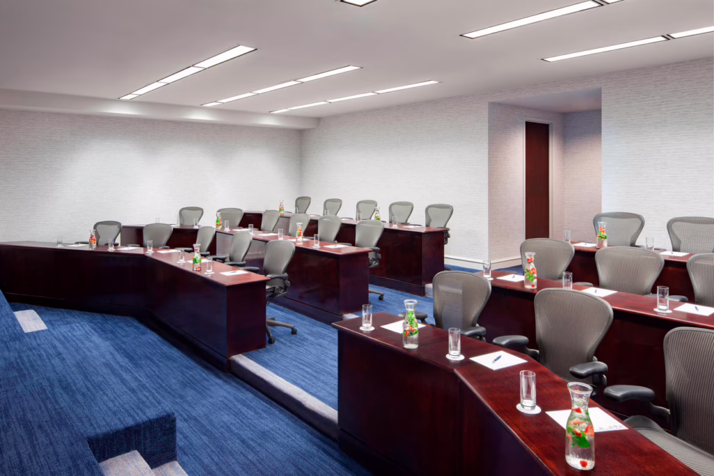 Modern conference room with rows of gray chairs, wooden desks, bottled water, glasses, and notepads on each table, under bright overhead lights.