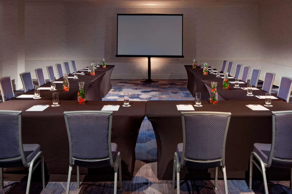 Conference room with U-shaped tables, gray chairs, water glasses, notepads, and a large blank projection screen at the front.