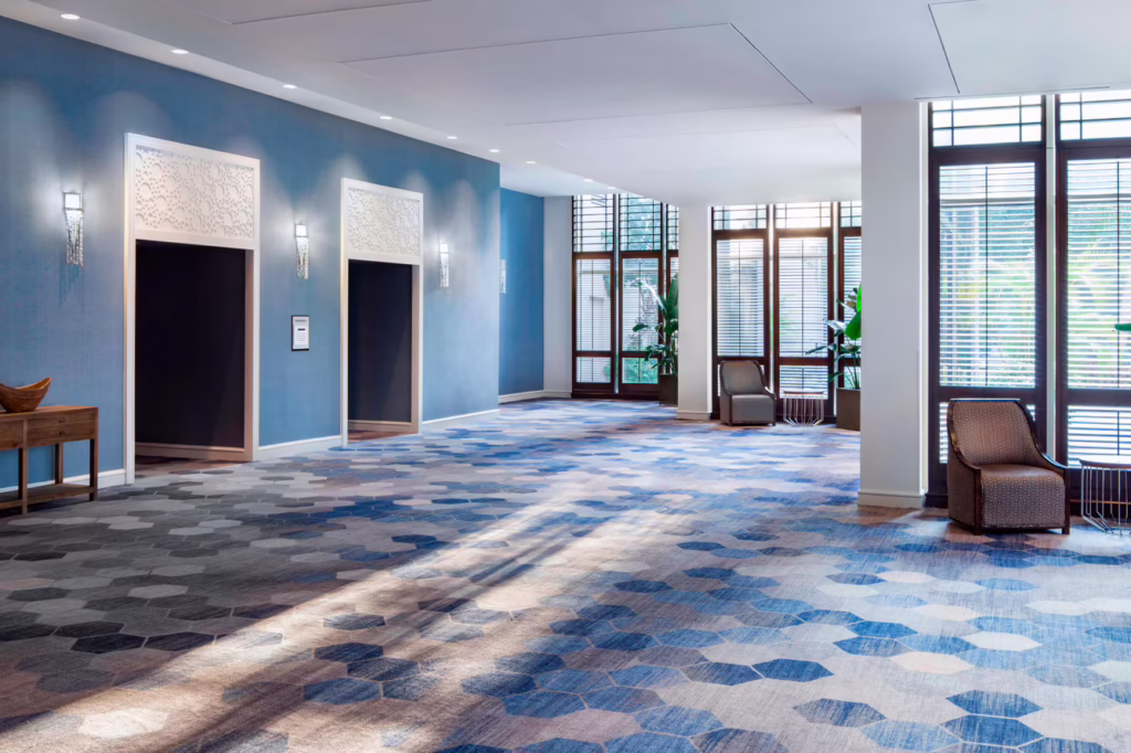 Elegant hotel lobby with blue patterned carpet, three elevators, large windows with blinds, sunlight streaming in, and modern armchairs by the windows.