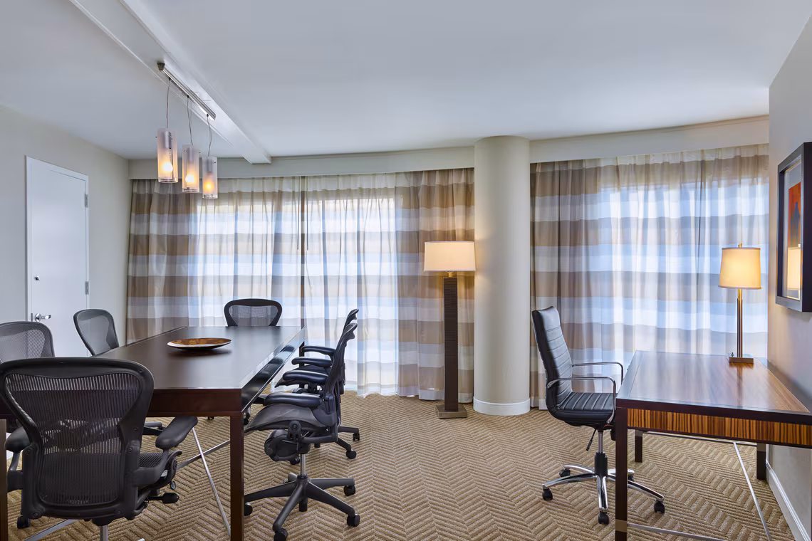 Modern conference room with a long table, six office chairs, a desk, floor lamp, and large windows with striped curtains letting in natural light.