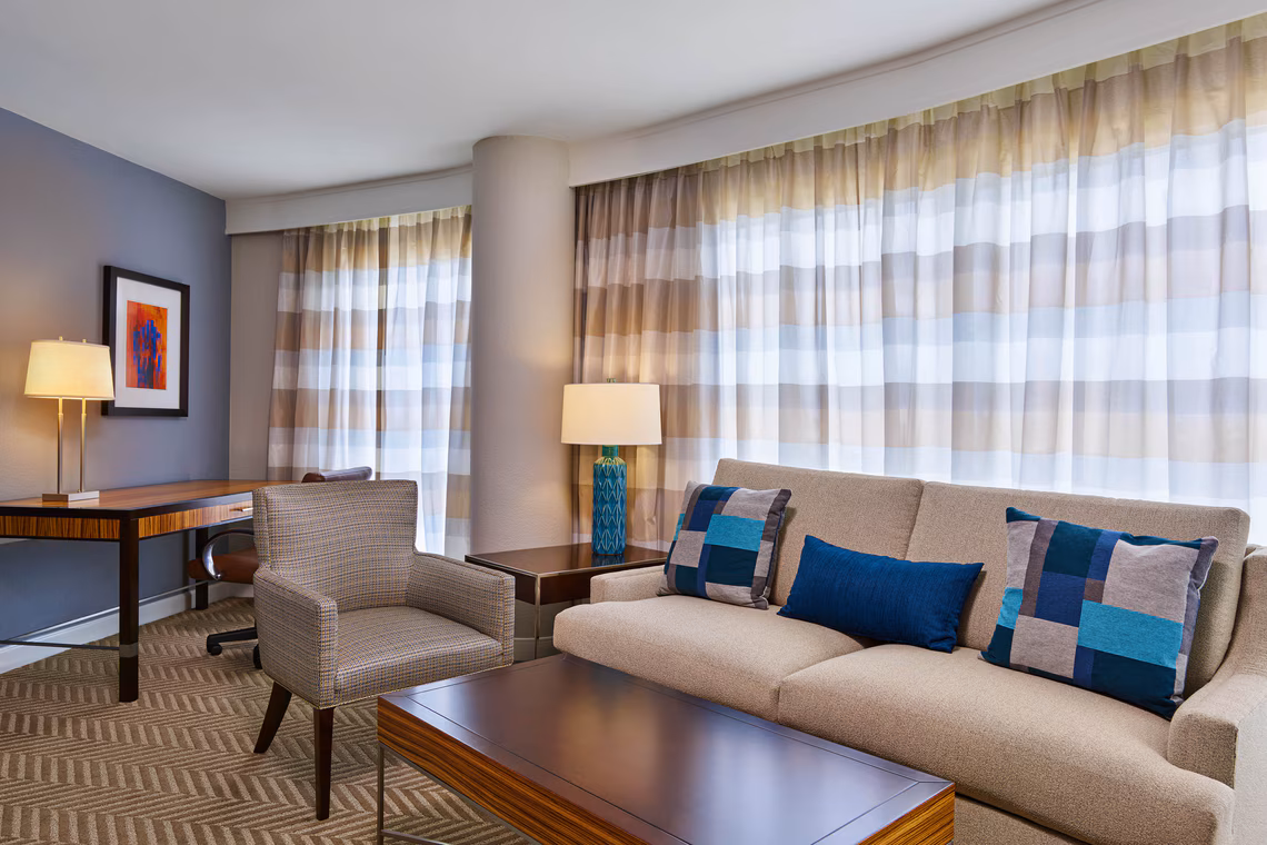 Modern hotel room with a beige sofa, colorful pillows, wooden desk, chairs, lamps, and large windows with striped curtains.