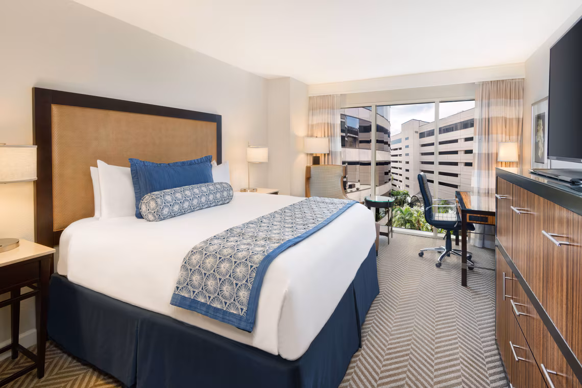 Hotel room with a large bed, blue and white bedding, desk, TV, and window showing buildings outside. Neutral decor with modern furnishings.