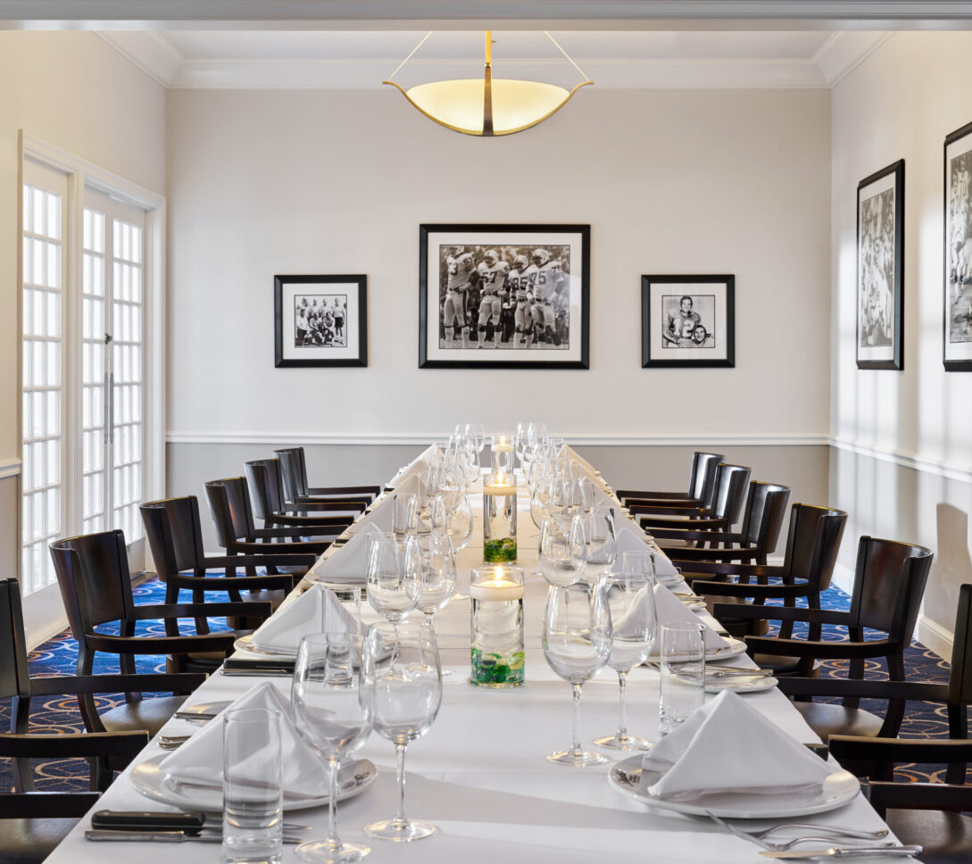 Elegant dining room with a long table set for a formal meal, surrounded by dark chairs, white walls, and black-and-white photos on the walls.