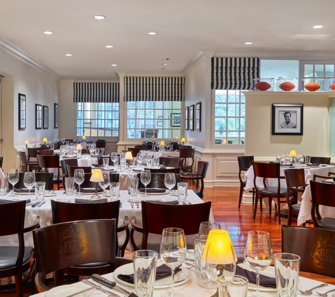 Elegant dining room with set tables, wooden chairs, striped curtains, framed photos, and footballs displayed on a shelf by the window.