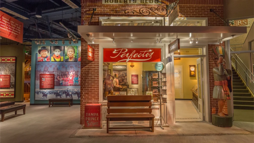 A museum exhibit of a vintage storefront with Perfectos sign, brick walls, a bench, mannequins, and historical displays inside and around it.