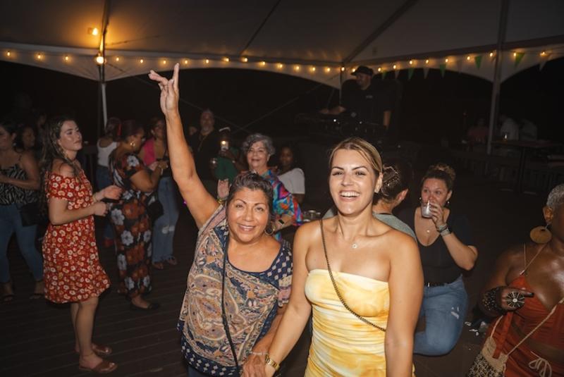 People dancing and smiling at an outdoor nighttime party under a tent with string lights. A woman in front raises her hand, celebrating.