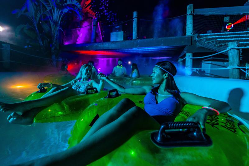 People in swimsuits relax on glowing tubes in a dimly lit indoor water park with colorful lights and mist.