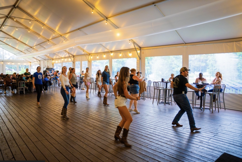 People in cowboy boots line dance on a wooden floor under a white tent, while others sit at tables in the background.