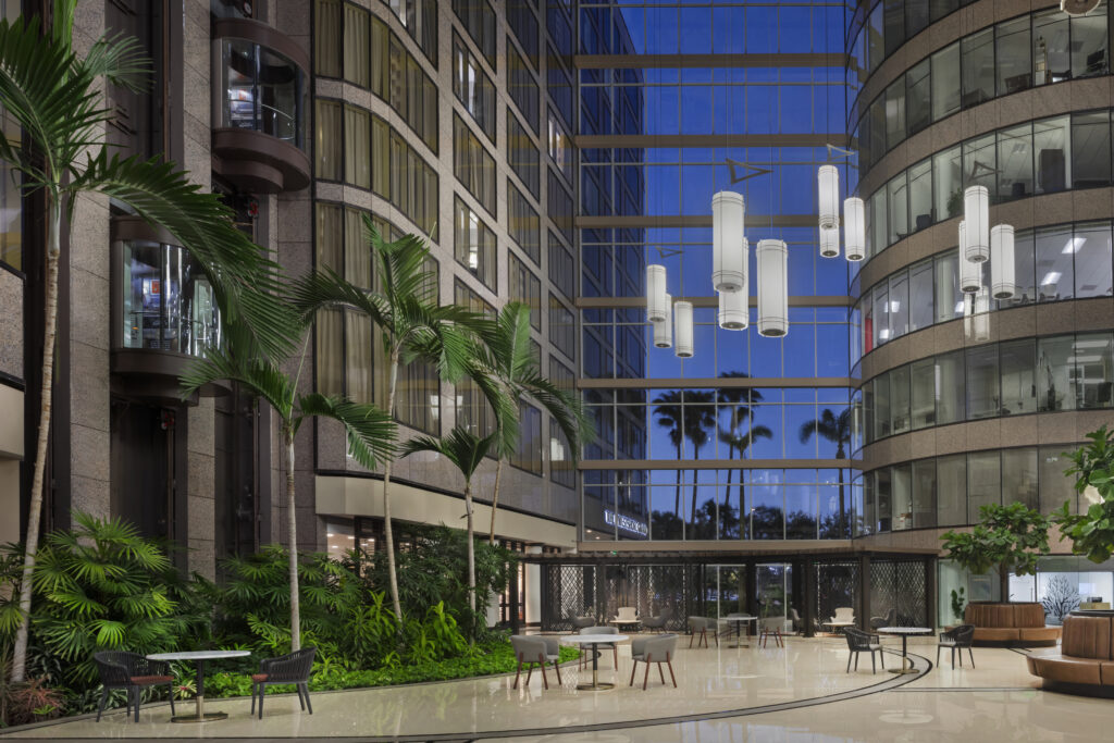 Modern lobby with tall glass windows, palm trees, hanging lights, indoor plants, tables, and chairs, reflecting an evening sky outside.