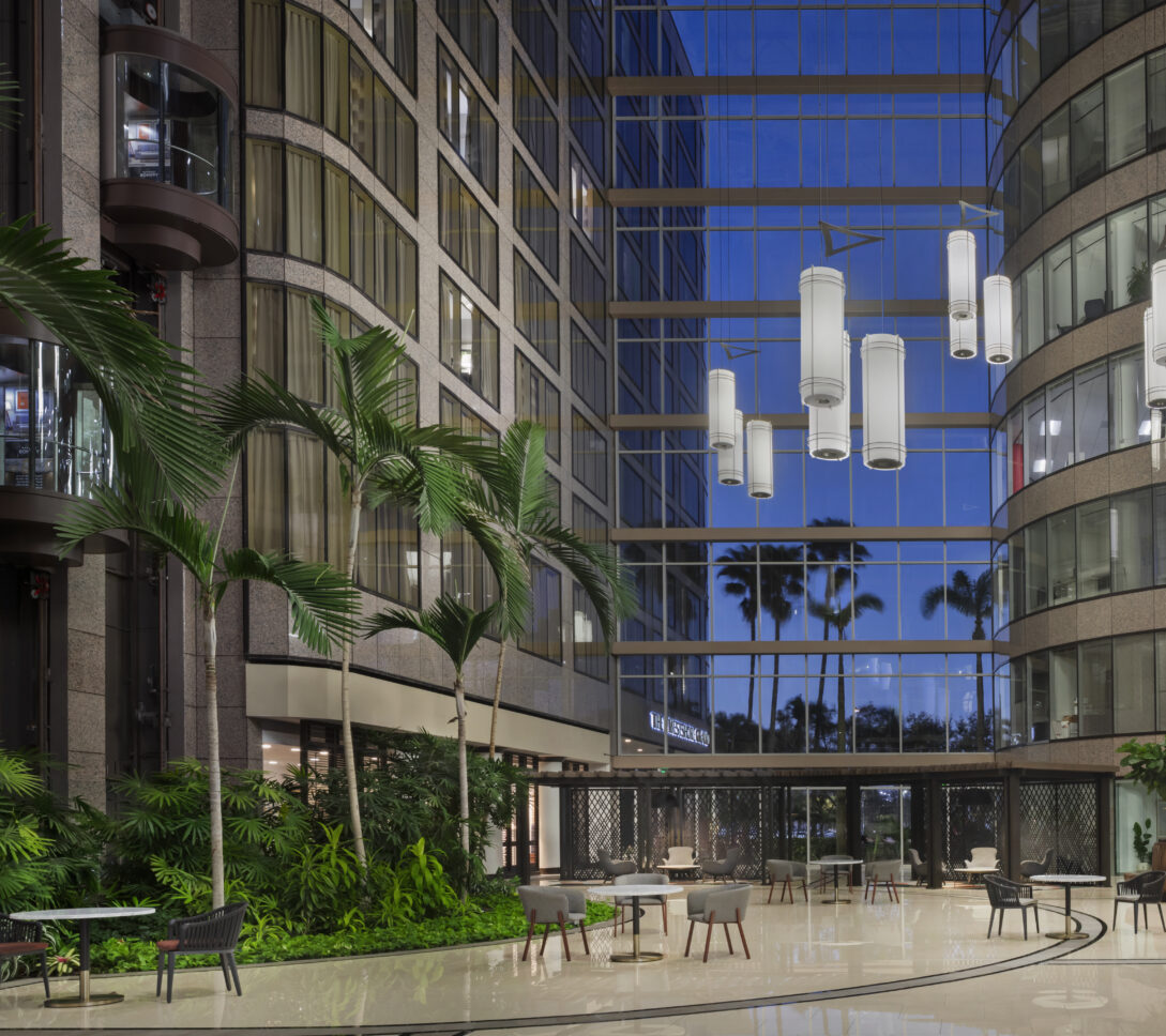 Modern lobby with tall glass windows, palm trees, hanging lights, indoor plants, tables, and chairs, reflecting an evening sky outside.