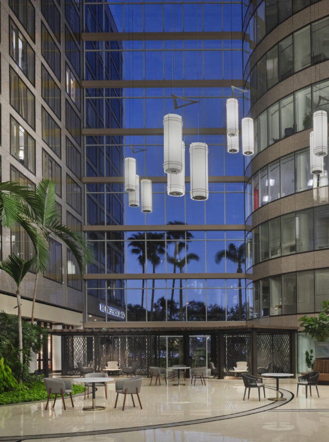 Modern lobby with tall glass windows, palm trees, hanging lights, indoor plants, tables, and chairs, reflecting an evening sky outside.