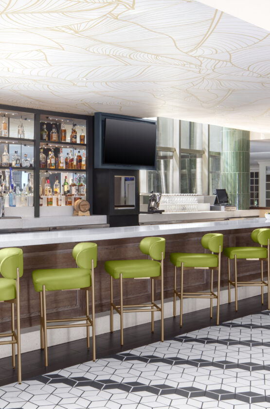Modern bar with green barstools, white counter, two TVs, and shelves of bottles. Geometric tile floor and colorful mural in the background.