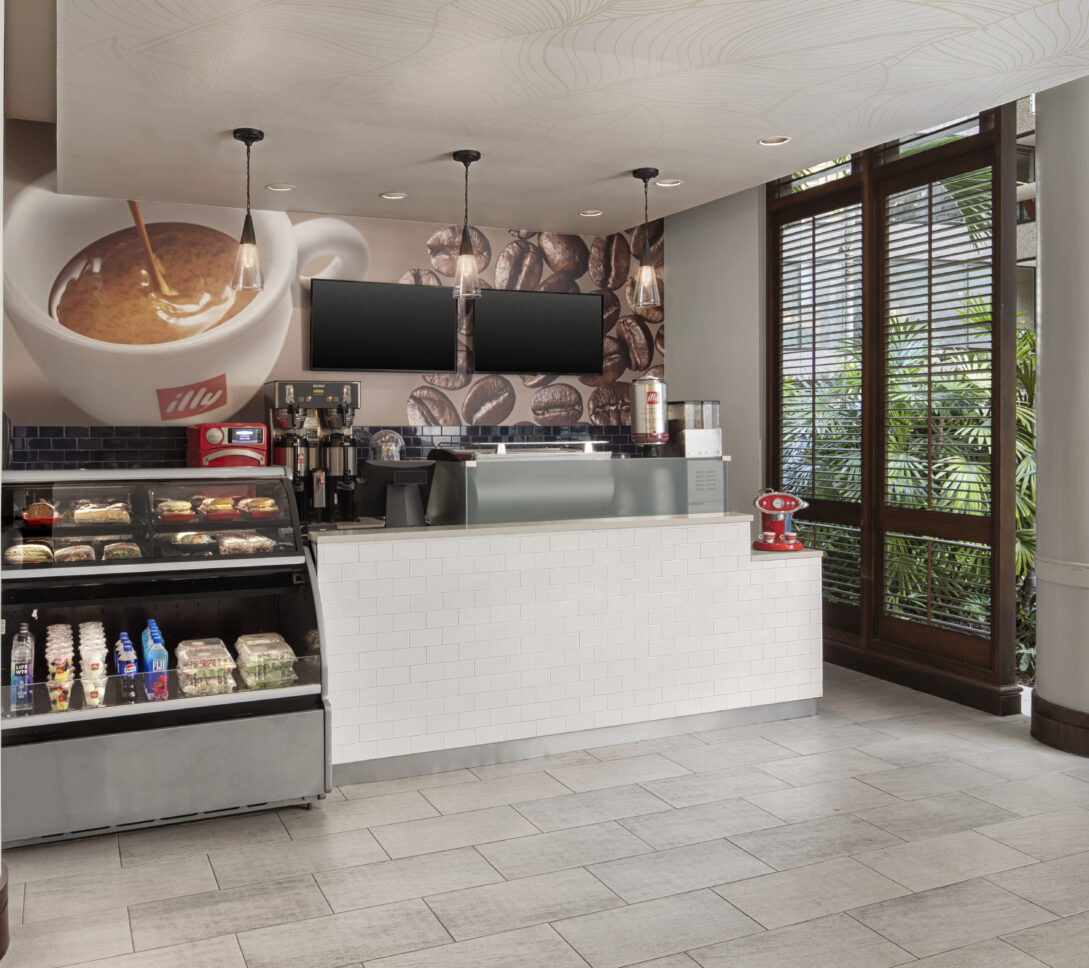 Modern coffee shop counter with display case, coffee machines, and two blank screens; large windows show green plants outside.