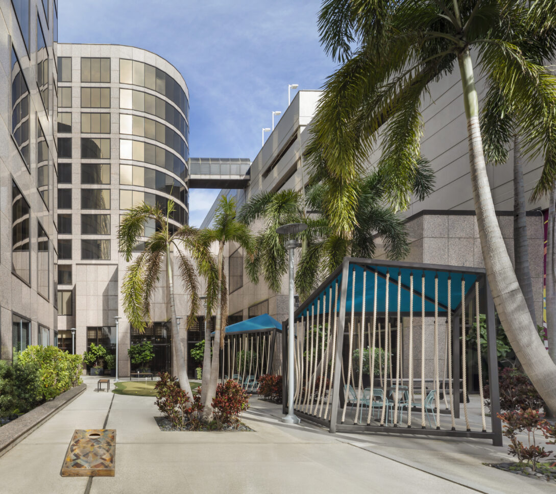 Modern courtyard with palm trees, shaded seating areas, colorful mural, and tall office buildings under a blue sky.