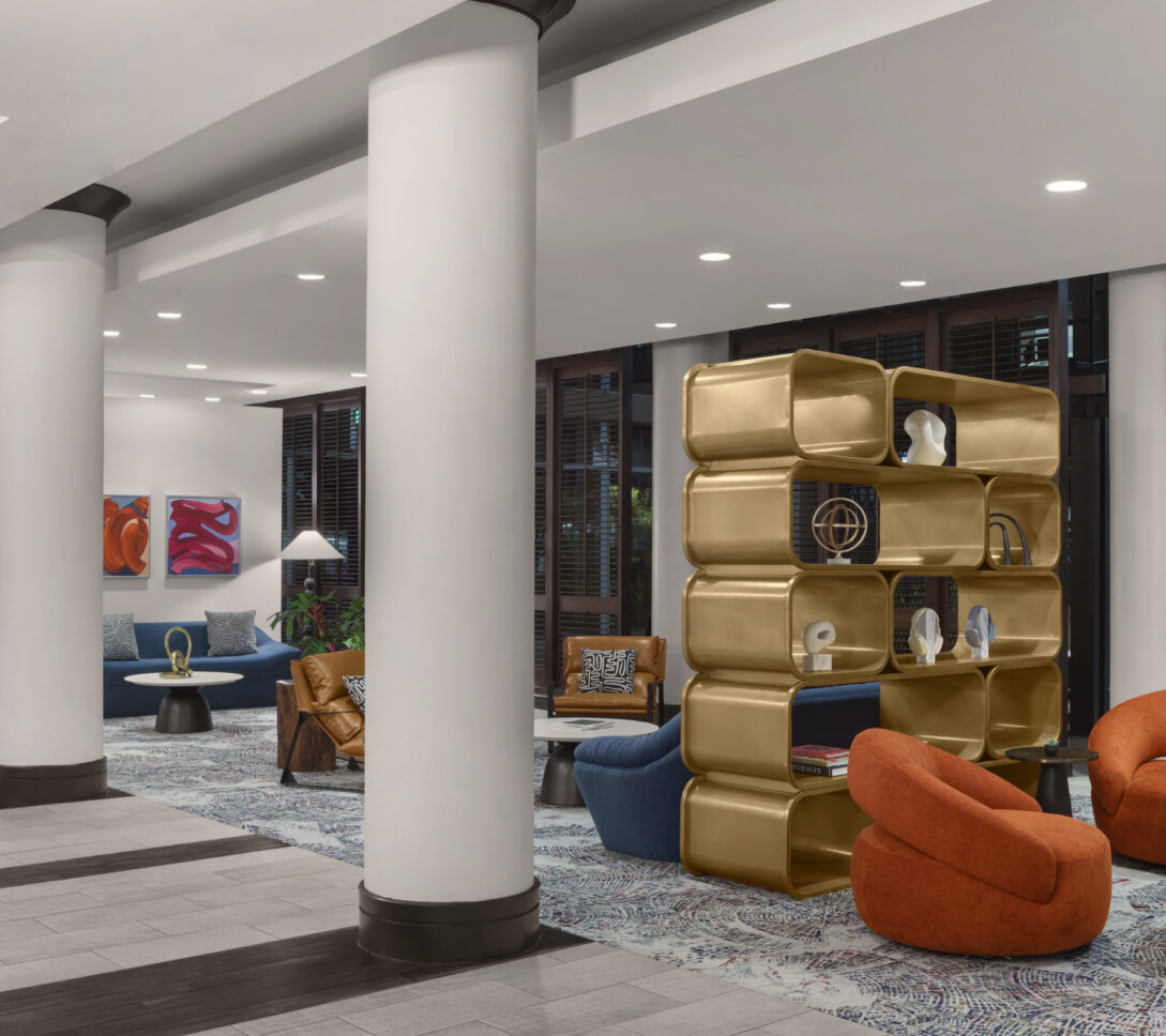 Modern hotel lobby with white columns, colorful chairs, a gold bookshelf, abstract art on the walls, and patterned carpet flooring.