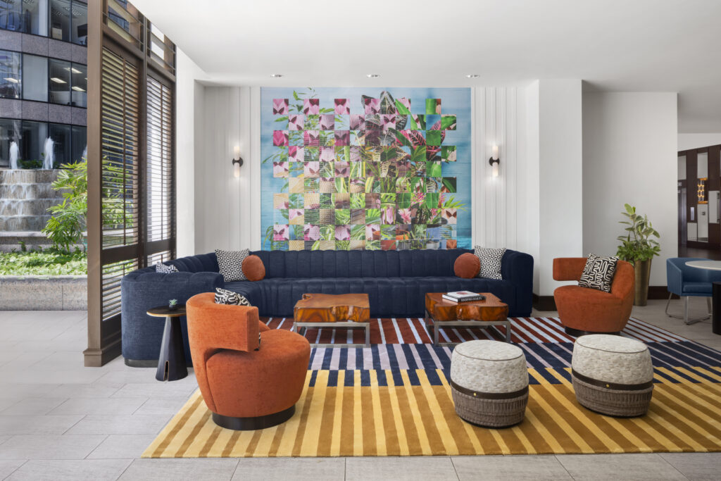 Modern hotel lobby with a blue sofa, orange chairs, striped rug, round ottomans, geometric wall art, and large window with blinds.