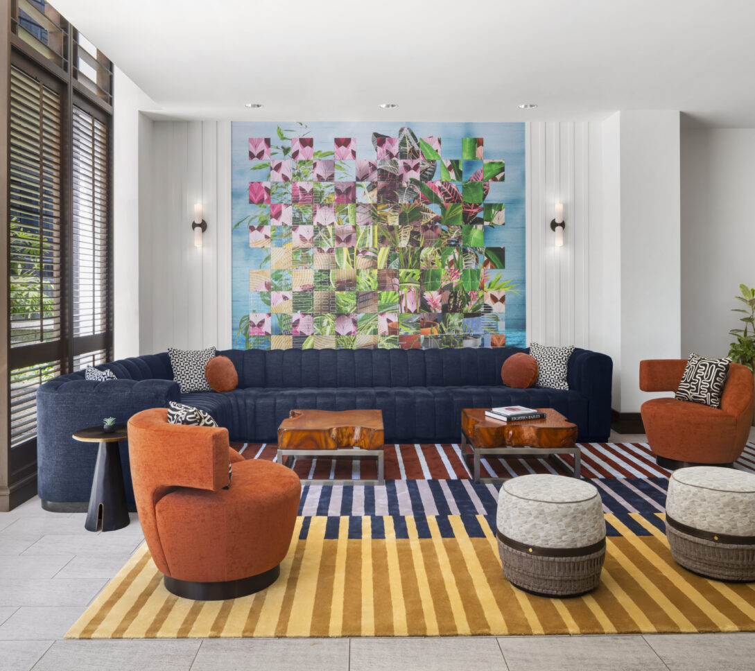 Modern hotel lobby with a blue sofa, orange chairs, striped rug, round ottomans, geometric wall art, and large window with blinds.
